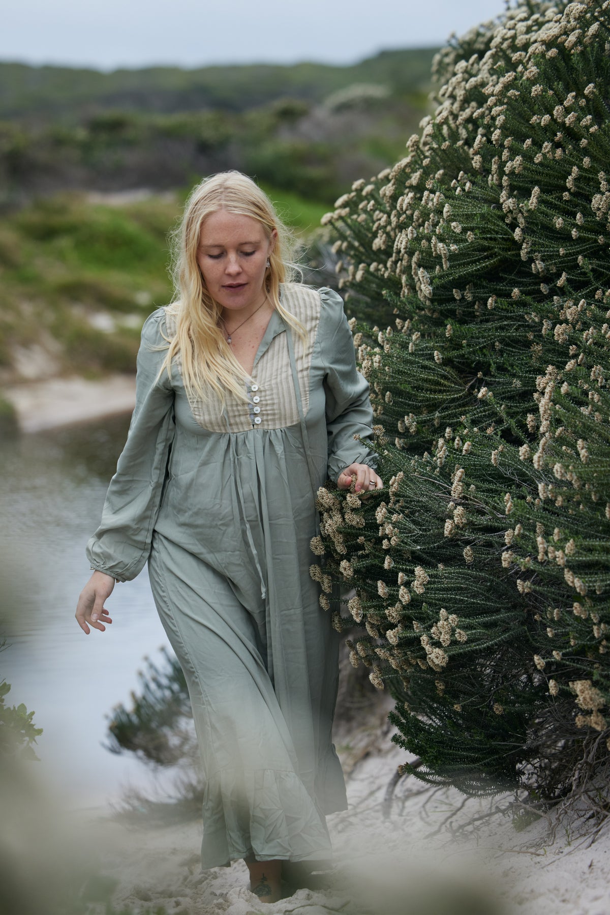 A woman with blonde hair wearing a sage green long-sleeve maxi dress walks outdoors next to a large green flowering bush.