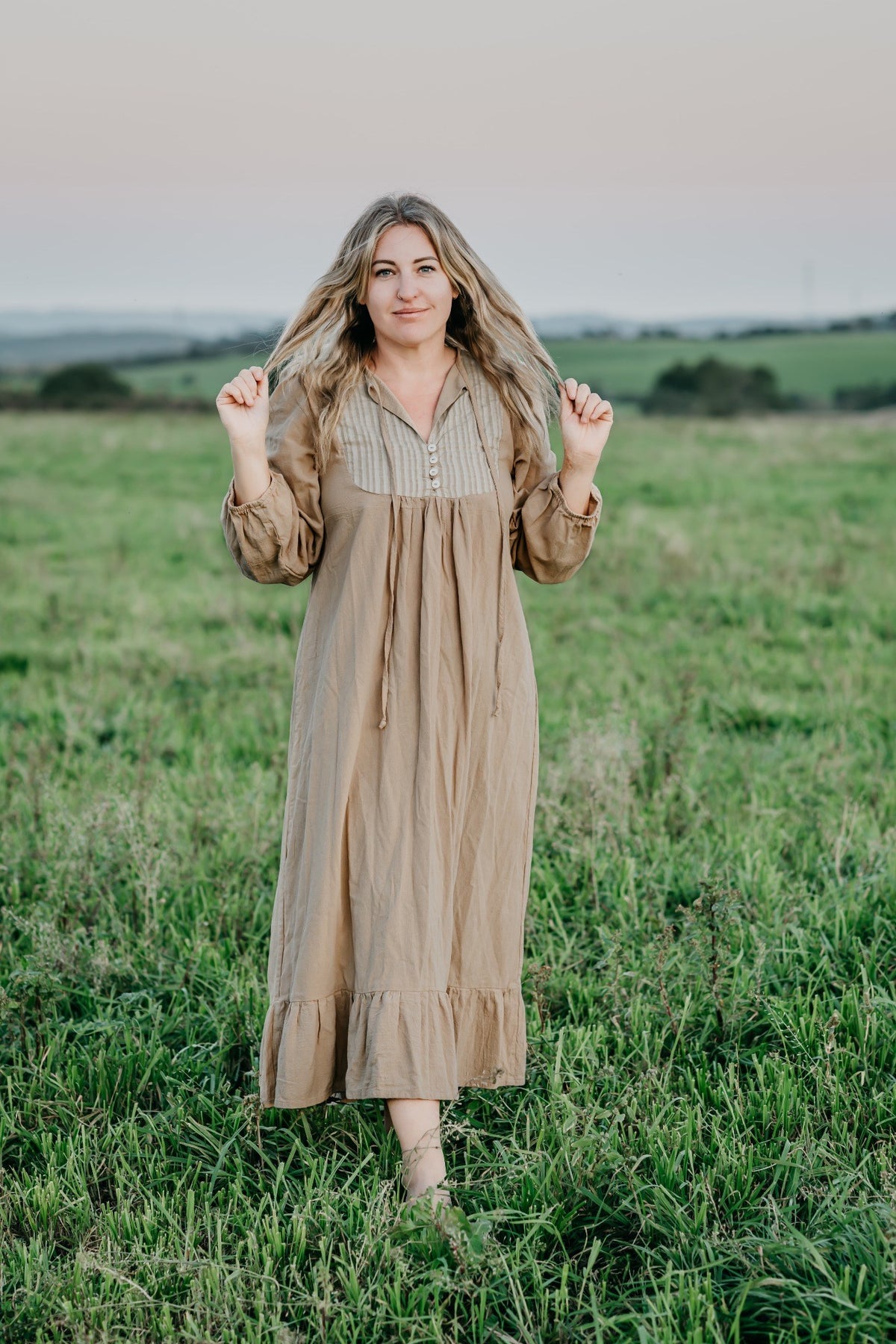 A woman stands in a grassy field wearing a long beige linen dress with a striped button-up bodice and long sleeves.