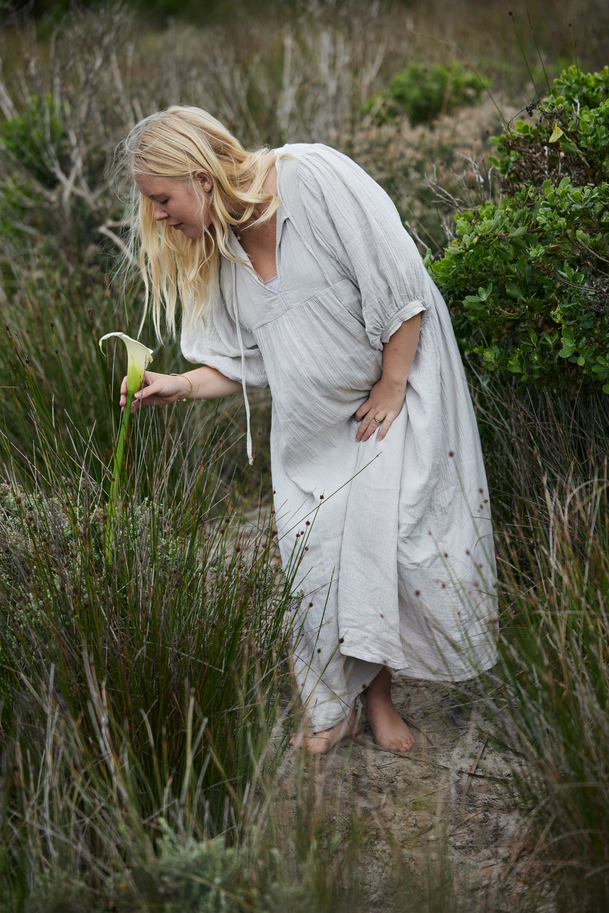 A woman with long blonde hair stands barefoot in a grassy outdoor setting, wearing a loose, light-coloured linen dress and holding a green and white calla lily plant.