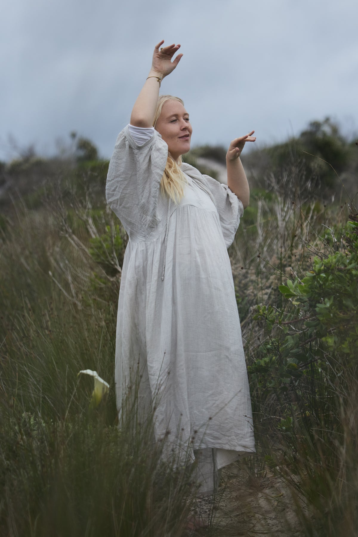 A woman wearing a long pale blue organic cotton dress stands outdoors in tall grassy terrain under an overcast sky, with her arms raised in an expressive pose.