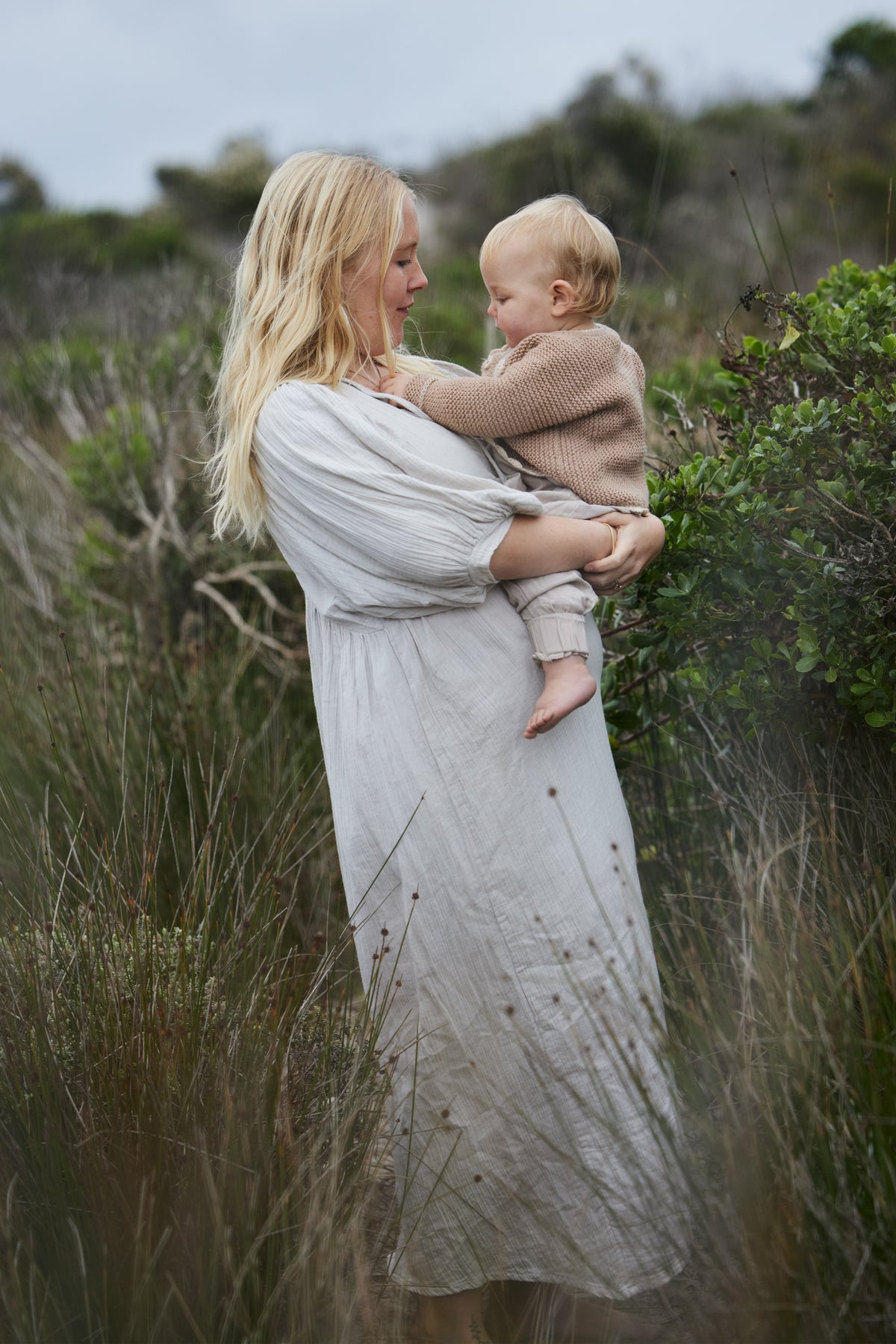 A woman in a long, loose pale blue dress holds a baby wearing a beige knit outfit, standing in a grassy outdoor field.