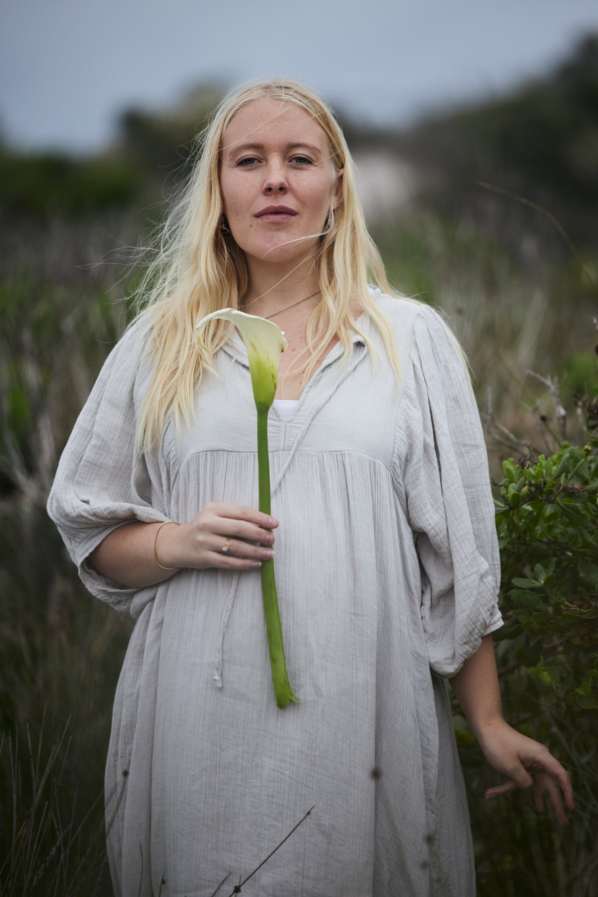A blonde woman wearing a loose pale blue, organic cotton dress stands outdoors in a grassy field, holding a white calla lily flower.