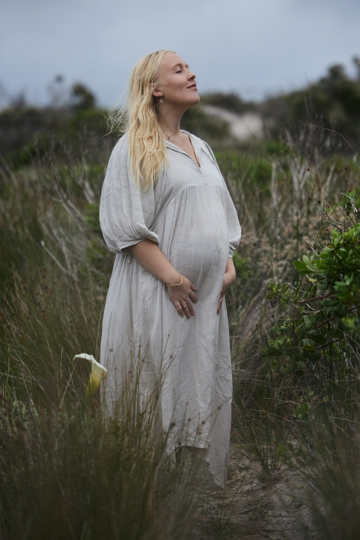 A pregnant woman with blonde hair stands outdoors in a grassy field, wearing a loose, light-coloured cotton dress. She is looking upward with her hands resting on her belly.
