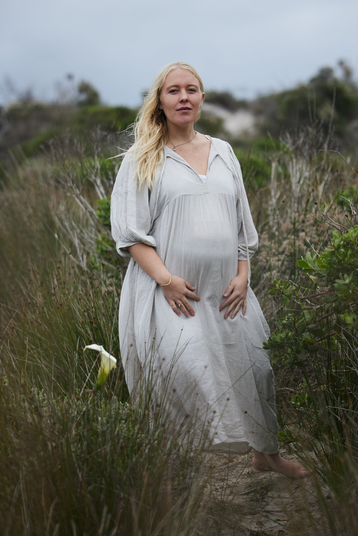 A pregnant woman stands barefoot in a grassy outdoor setting, wearing a loose, long-sleeve pale blue, flowing dress.