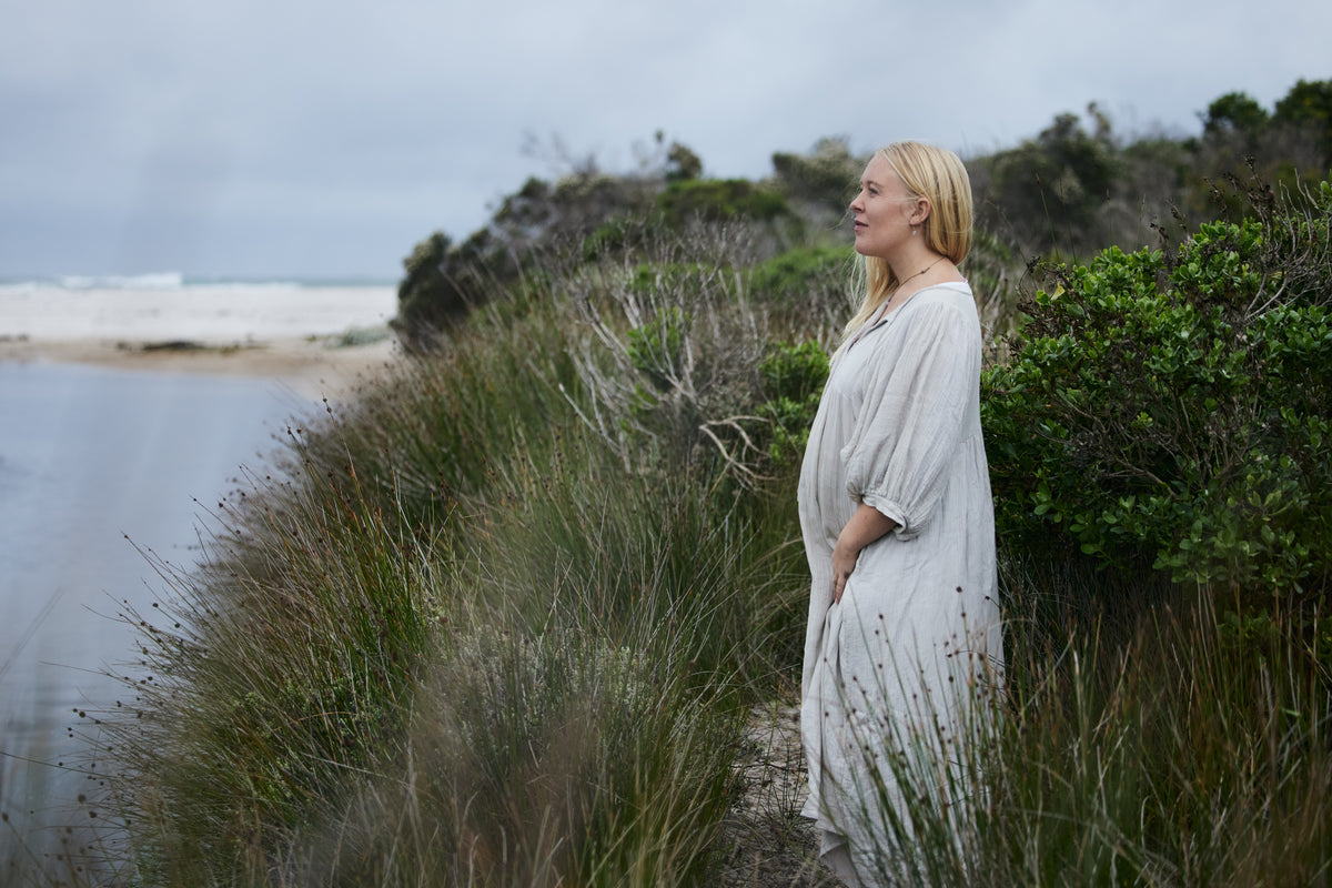 A pregnant woman with blonde hair stands in profile on a grassy coastal path, wearing a loose, long-sleeve dress. The background features green beach grass, shrubs, and an overcast ocean sky.