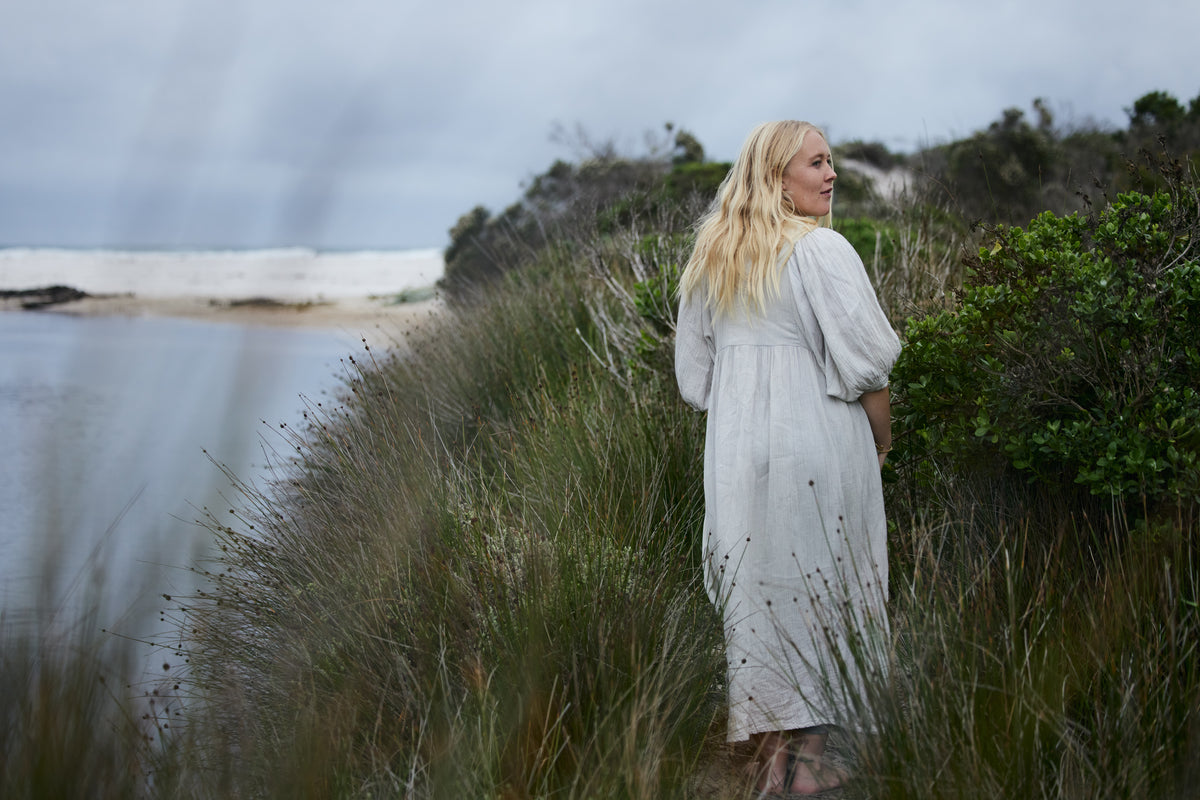 A woman with long blonde hair stands on a grassy coastal dune wearing a loose, light-colored maxi dress, looking out towards the ocean under an overcast sky.