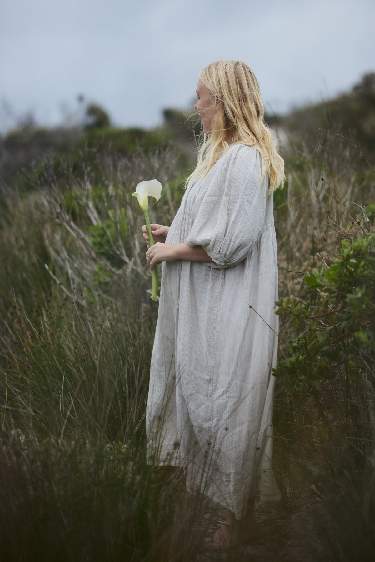 A woman with long blonde hair stands in a grassy outdoor field wearing a loose, light-colored maxi dress and holding a white calla lily flower.