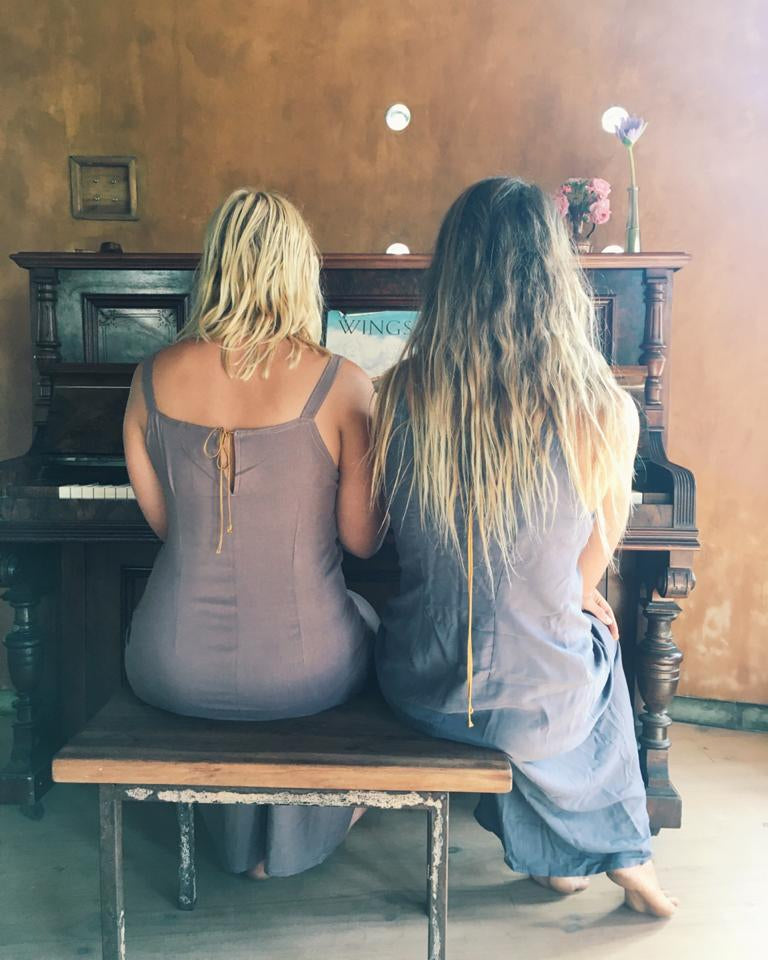 Two women sitting on a bench in front of a dark wooden piano, viewed from behind. The woman on the left wears a sleeveless taupe dress, and the woman on the right wears a long-sleeved dusty blue dress.
