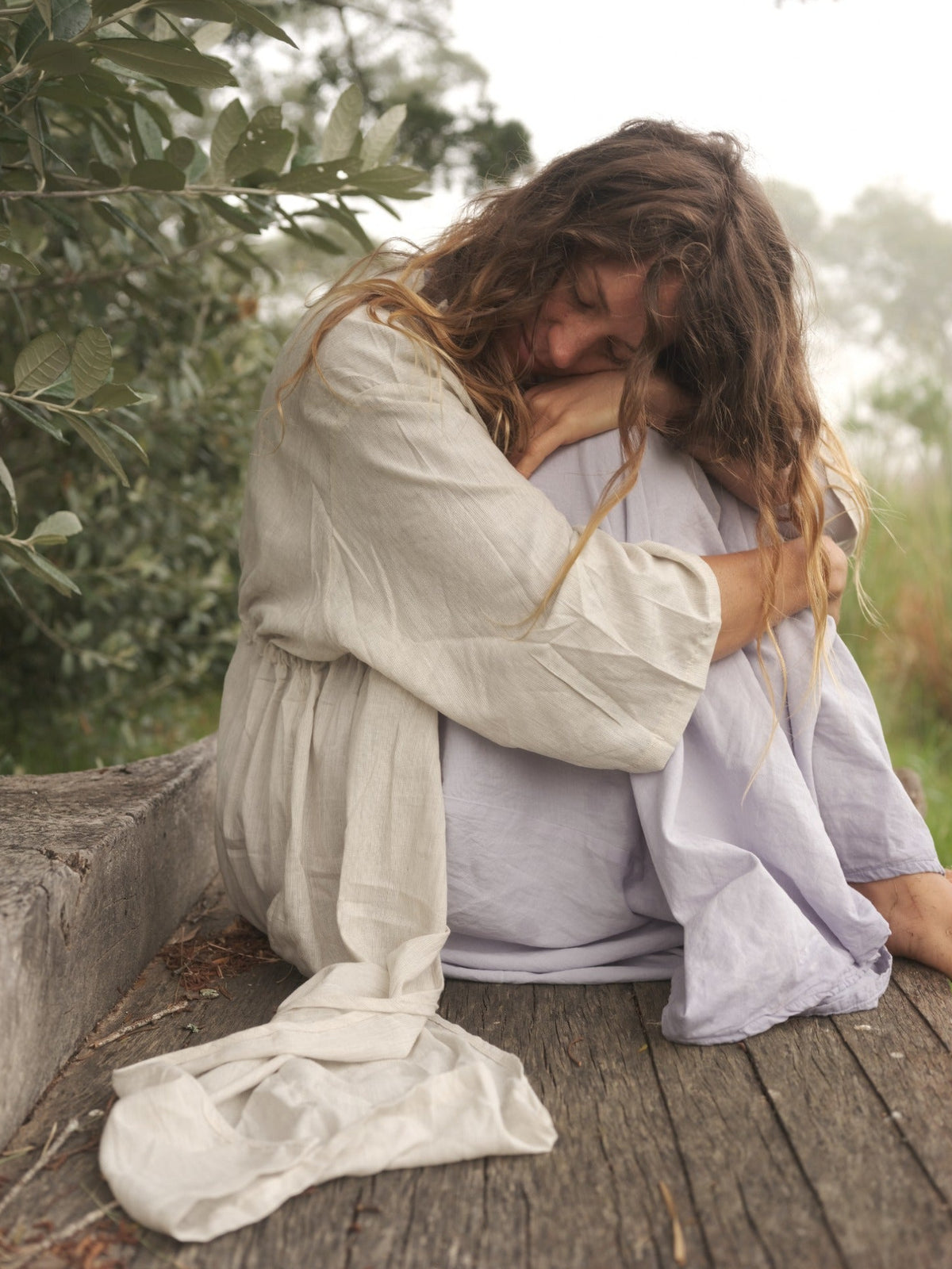 A woman with long brown hair sits curled up on a weathered wooden bench outdoors, wearing a loose light beige kimono and a pale blue dress. Green foliage is visible in the background.