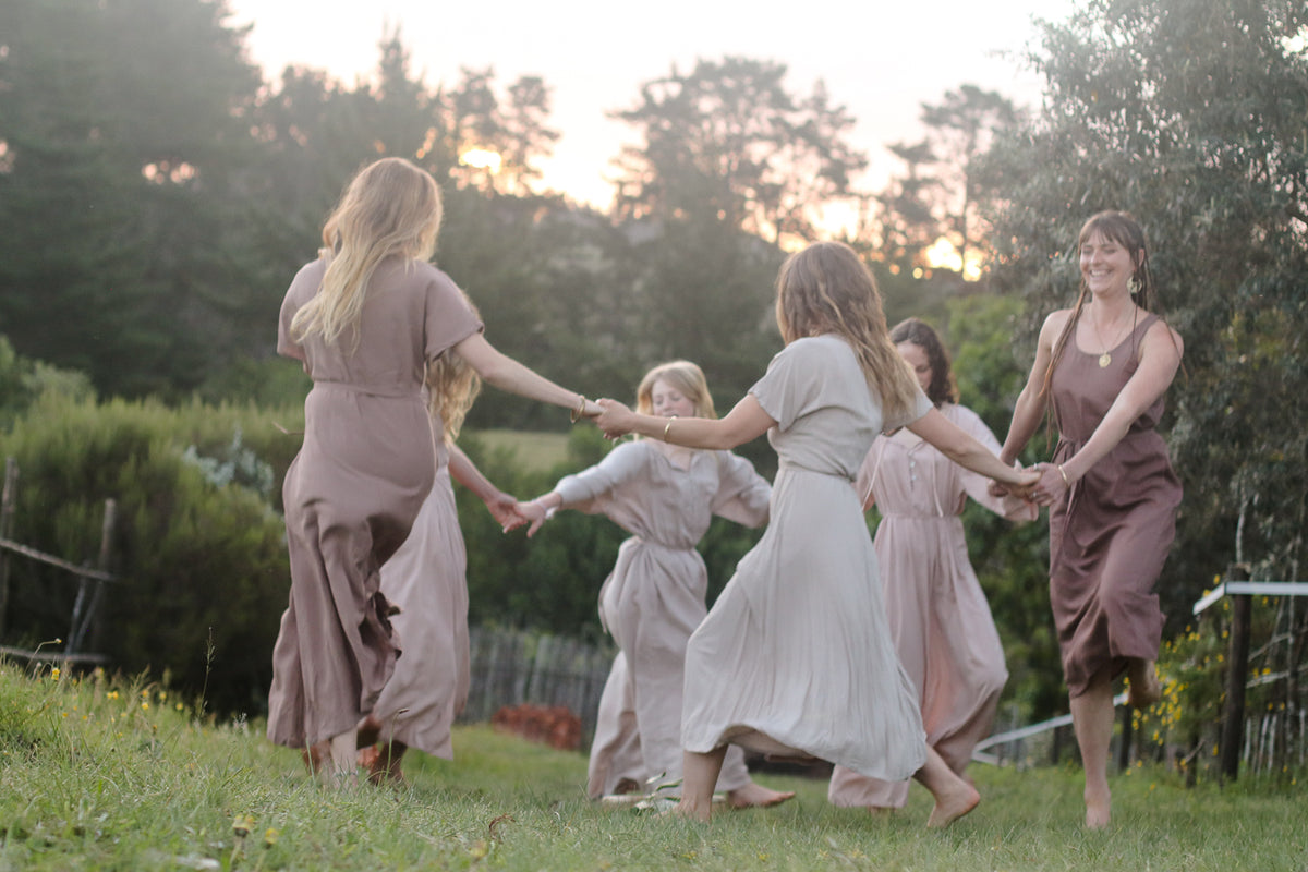 A group of women in matching muted dresses holding hands and dancing in a grassy outdoor garden at sunset.