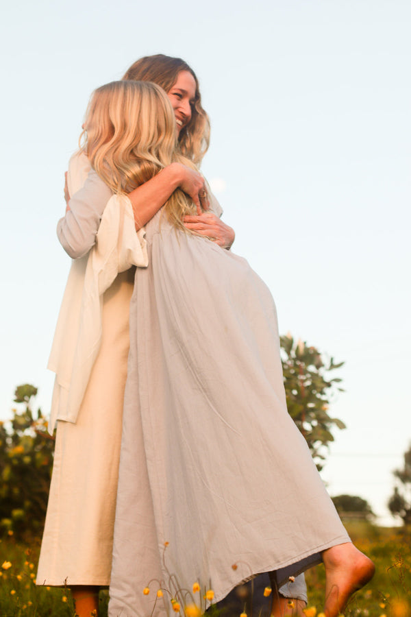 Two women embracing outdoors in a field of small yellow flowers, wearing long flowing linen-like dresses in soft neutral tones of beige, cream, and pale blue.