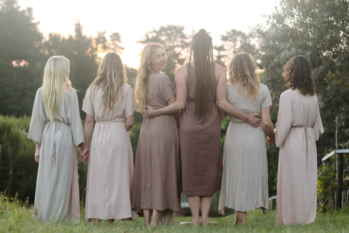 Six women standing outdoors at sunset, viewed from behind, wearing matching long draped maxi dresses in soft neutral tones of beige, taupe, blush, and dusty rose.
