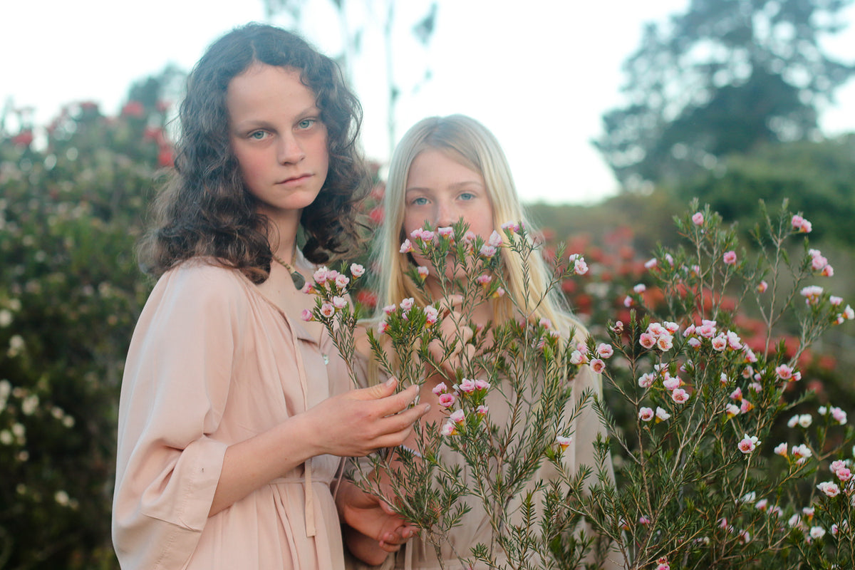 Two young girls standing outdoors in a garden among pink and white flowers, wearing soft light pink and beige linen-like dresses.
