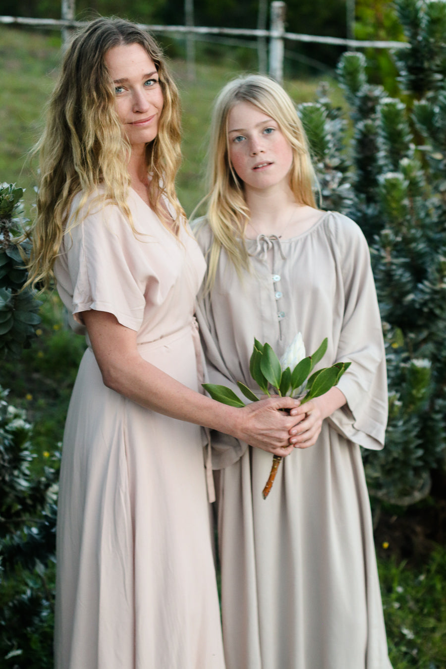 A woman and her daughter stand outdoors in matching light beige long-sleeve dresses, with the woman holding the girl's hands and the girl holding a small bouquet of green leaves and white flowers. The background shows green garden foliage.