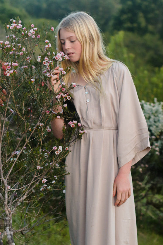 A young woman in a long beige linen dresses standing in a grassy garden, smelling small pink flowers.