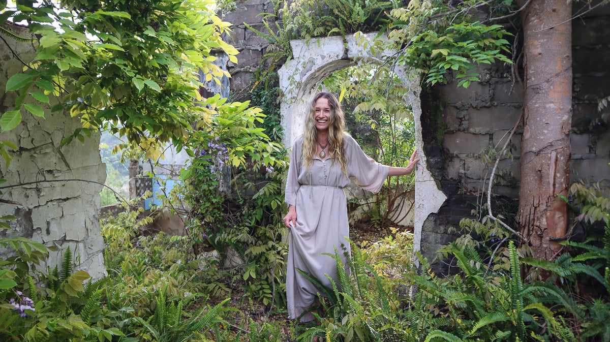 A woman wearing a long, loose beige dress stands in a lush, overgrown stone ruin garden, holding the side of a white archway.