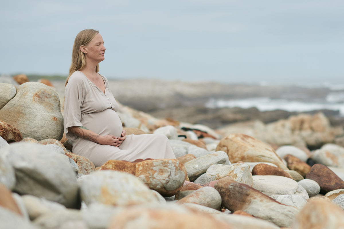 A pregnant woman in a light beige maternity dress sits on large rocks by the ocean, looking out toward the water under an overcast sky.