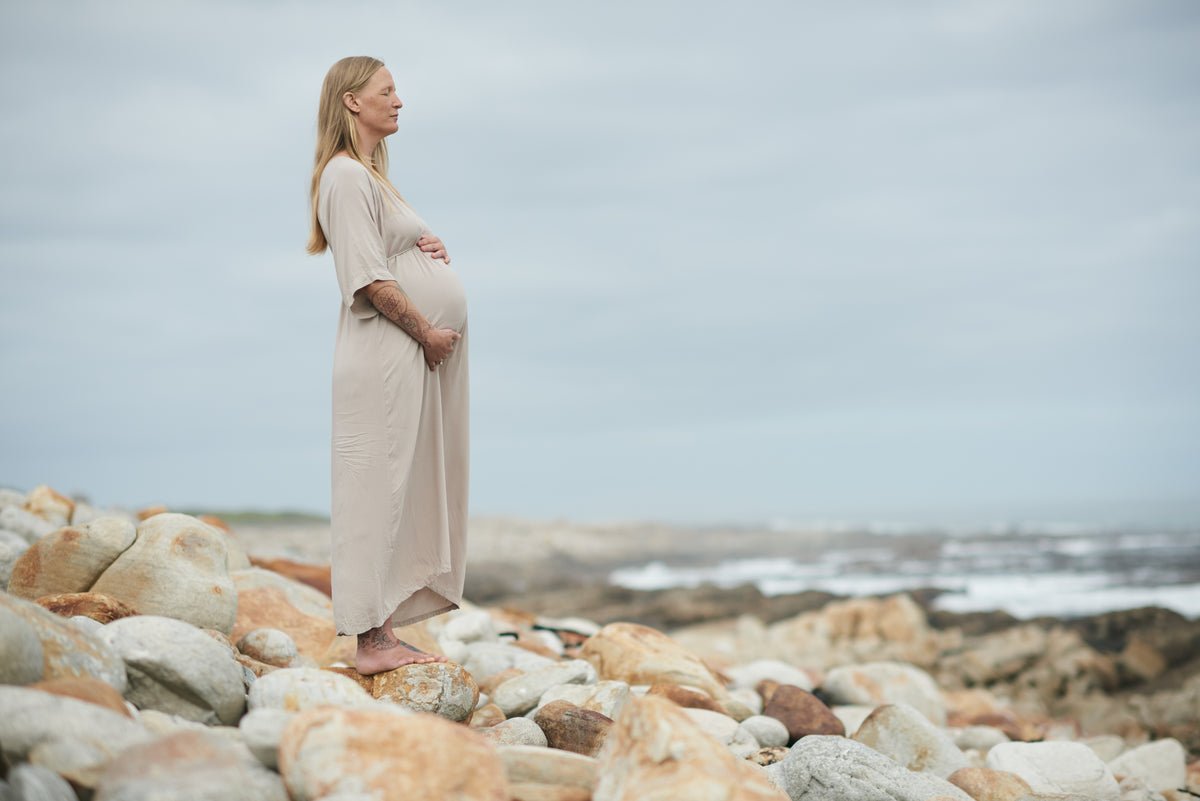 A pregnant woman in a long beige maxi dress stands barefoot on a rocky beach, holding her belly in profile under an overcast sky.
