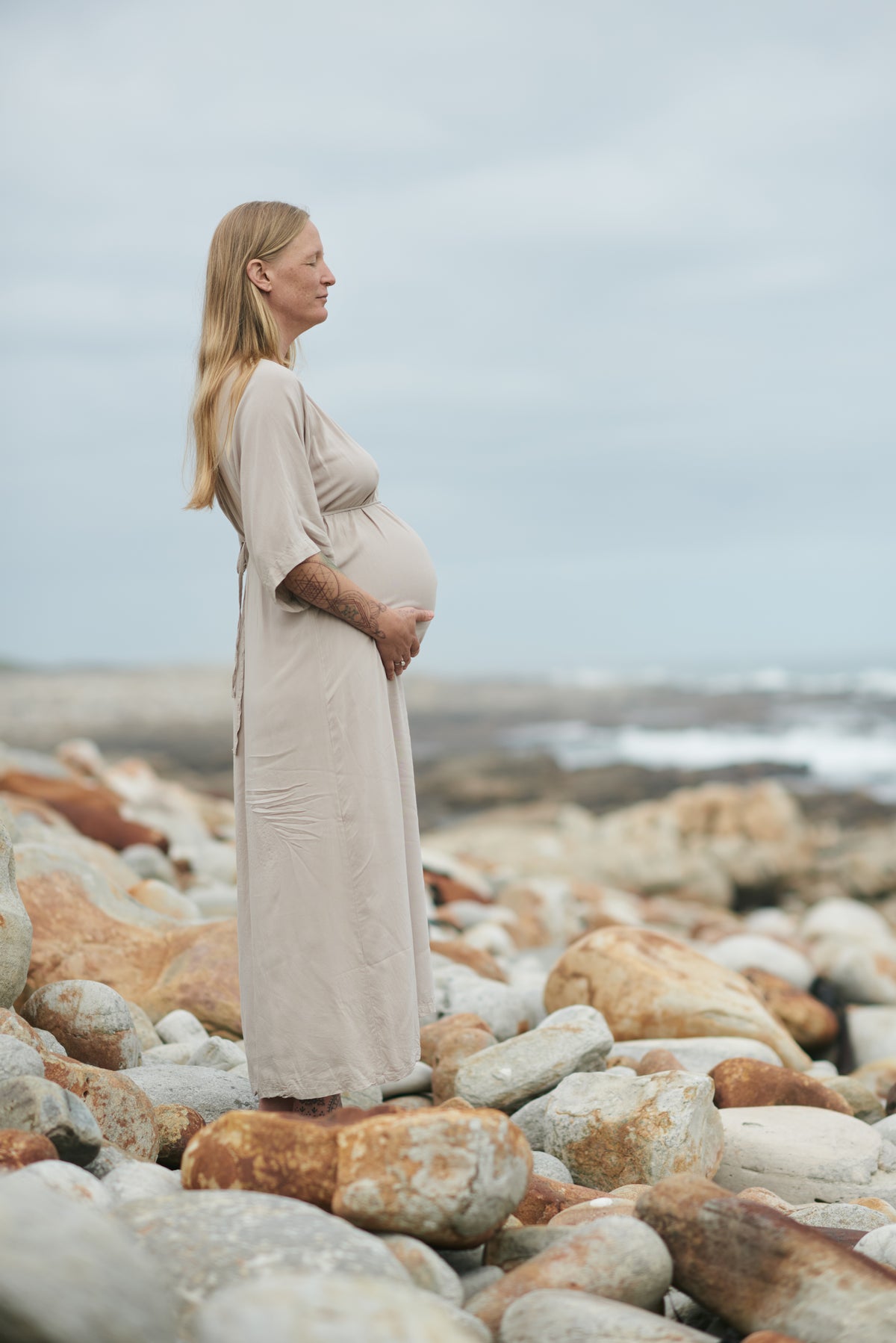 A pregnant woman stands in profile on a rocky beach, wearing a long, loose beige maternity dress. The background features a soft-focus ocean and cloudy sky.