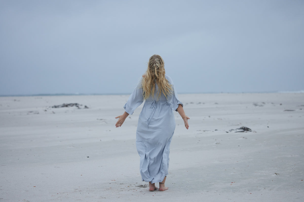 A woman with long blonde hair walks barefoot on a wide, pale sandy beach, seen from behind, wearing a loose light blue maxi dress with her arms outstretched. The sky is overcast and the horizon is low in the frame.