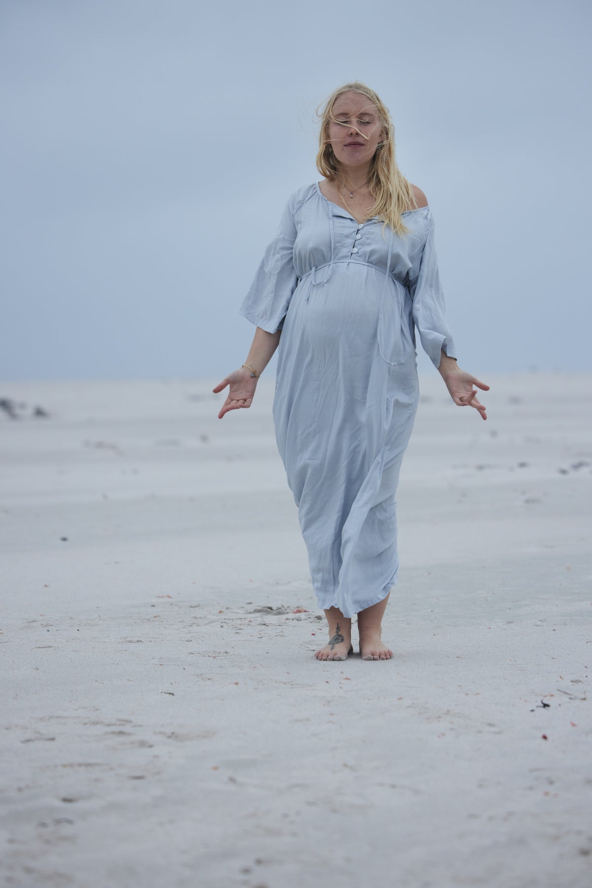 A pregnant woman walking barefoot on a sandy beach wearing a light blue off-the-shoulder maxi dress with long sleeves.