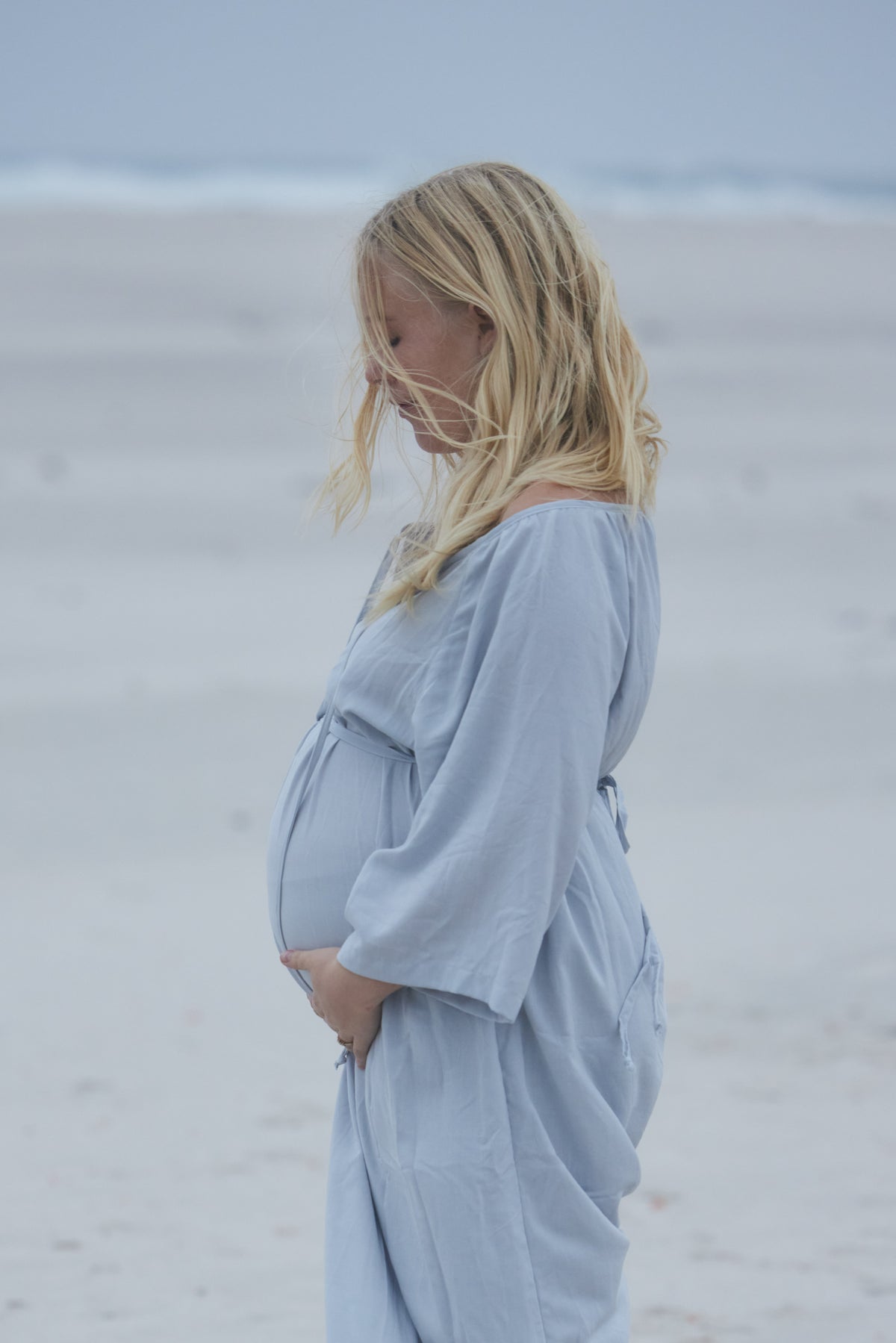 A pregnant woman with blonde hair stands in profile on a sandy beach, wearing a light blue long-sleeve maternity robe.