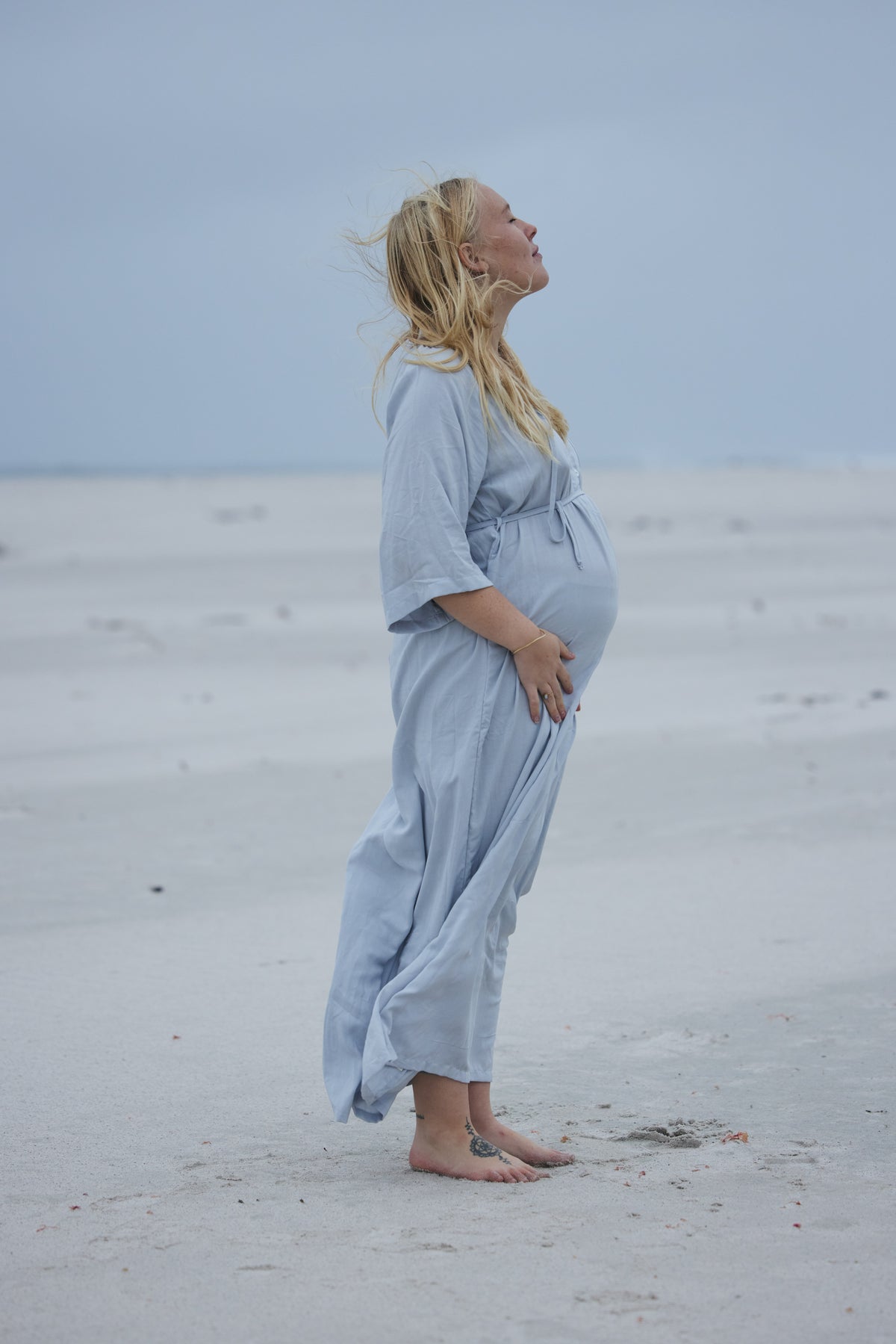 A pregnant woman stands barefoot on a sandy beach wearing a light blue maternity maxi dress, looking upward against a cloudy sky.