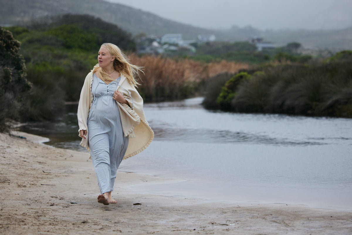 A pregnant woman walks barefoot along a sandy beach next to a calm river, wearing a light blue maternity jumpsuit and a cream-colored shawl.