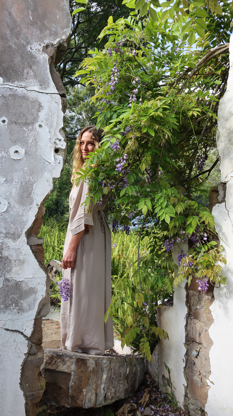 A woman wearing a long, loose beige dress stands in a narrow stone passage framed by white walls, with lush green foliage and purple flowers growing along the right side.