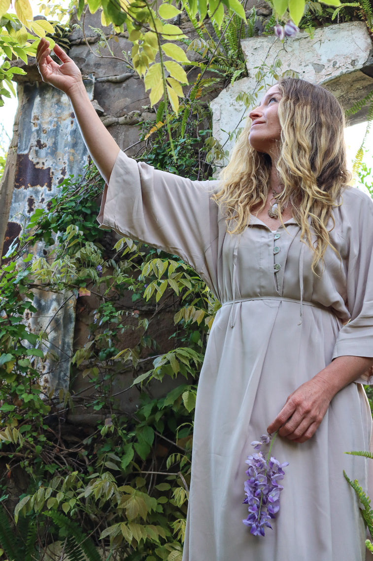 A woman in a light beige dress stands outdoors among green foliage and stone walls, reaching toward purple flowers while holding a small bunch.