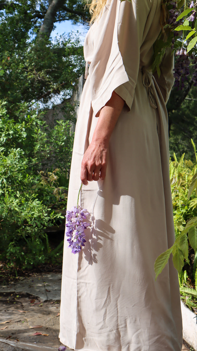 A woman outdoors in a garden wearing a long cream linen dress, holding a bunch of purple wisteria flowers. The dress has a tie-waist and wide sleeves.