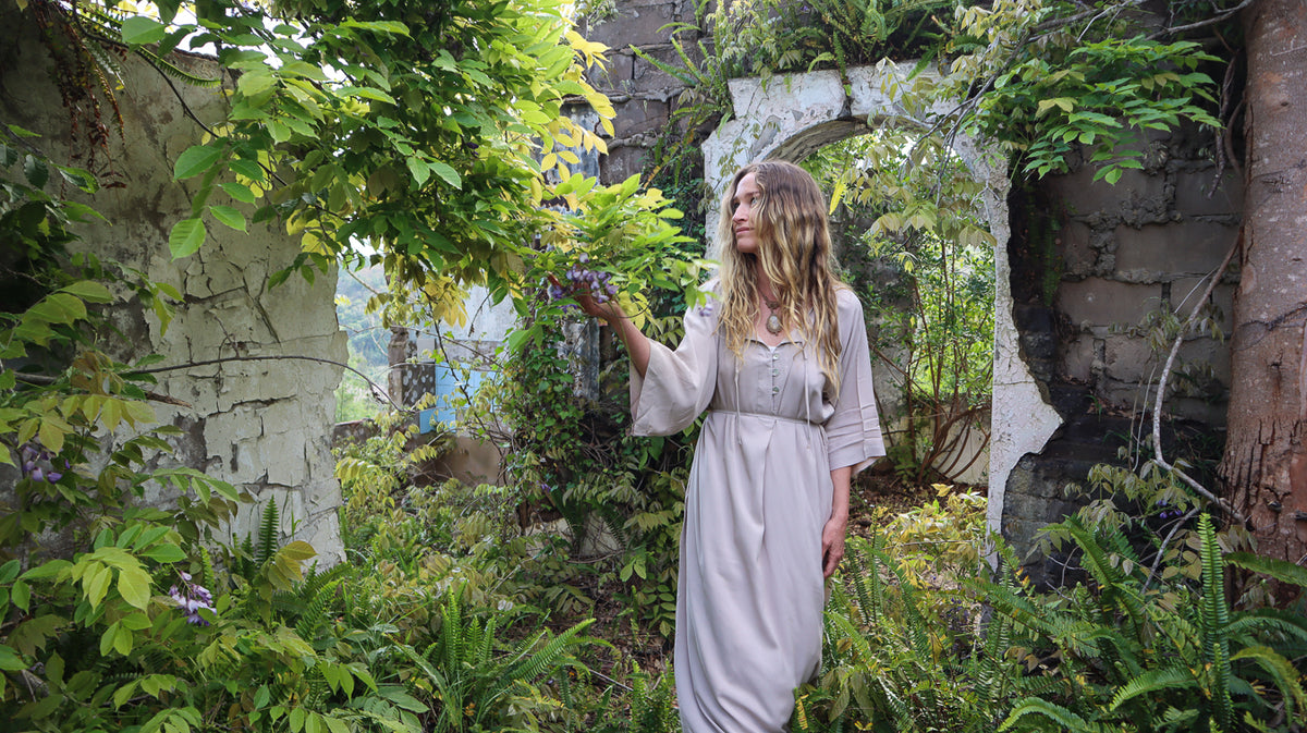 A woman wearing a light beige maxi dress stands in a lush, overgrown stone ruin garden, reaching toward green leaves.