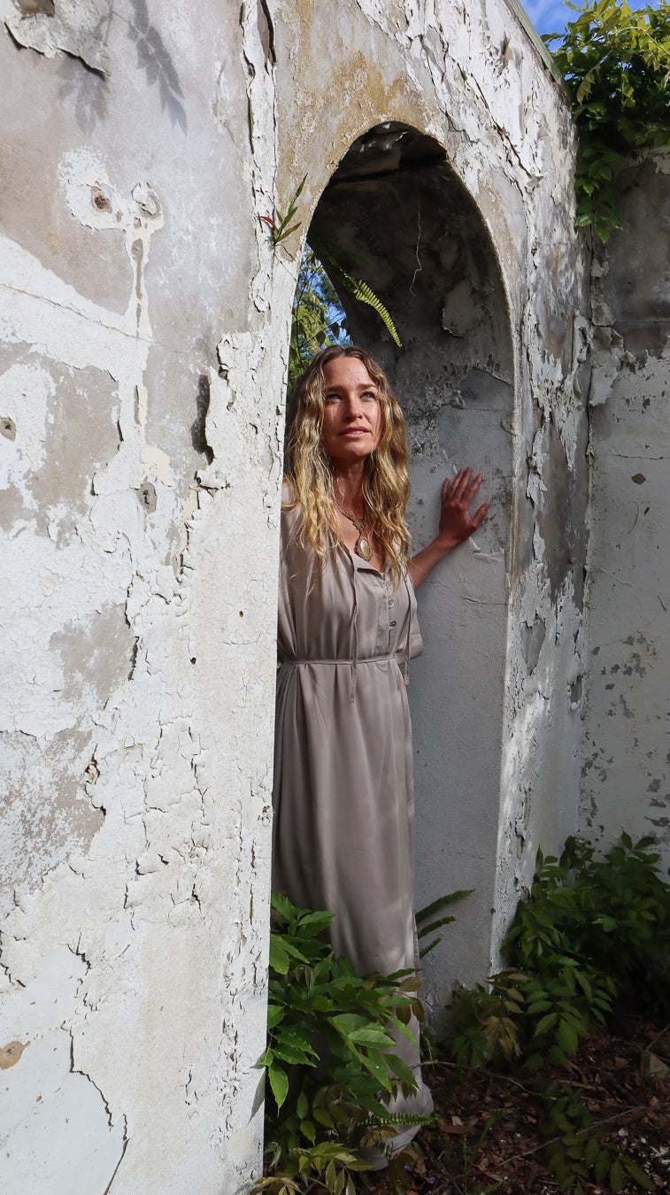 A woman with blonde hair wearing a light taupe maxi dress stands inside a weathered, peeling-paint archway doorway surrounded by green foliage.