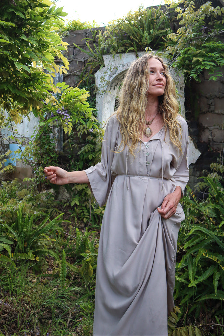 A woman with long blonde hair wearing a light beige linen dress stands in a lush green garden setting.