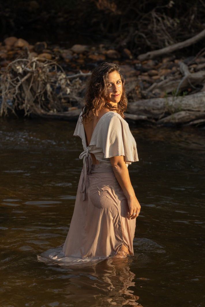 A woman stands in shallow water at a riverbank, wearing a flowing light beige dress with short flutter sleeves and a back tie. Fallen trees and roots line the riverbed behind her.