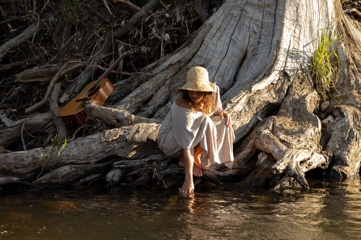 A woman wearing a straw hat and a light beige dress sits barefoot on a large tree root beside shallow water, with a guitar resting nearby in a natural outdoor setting.