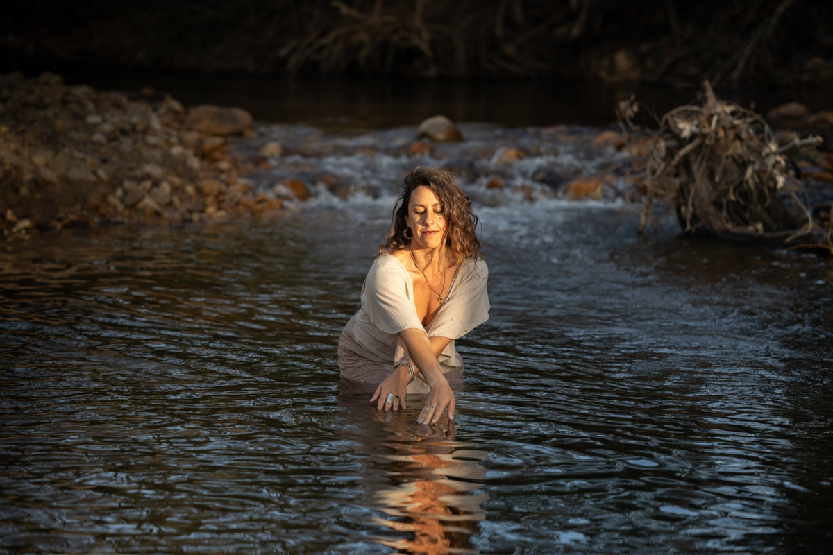 A woman stands knee-deep in a shallow river or stream, wearing a light beige dress. The background features dark, rocky banks and natural water ripples under warm, low-angle sunlight.
