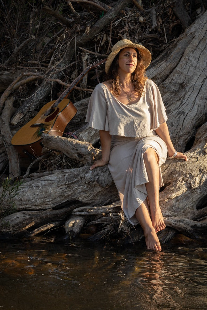 A woman wearing a beige bohemian-style dress  with a straw hat sits barefoot on a large log by a river, with an acoustic guitar resting nearby.