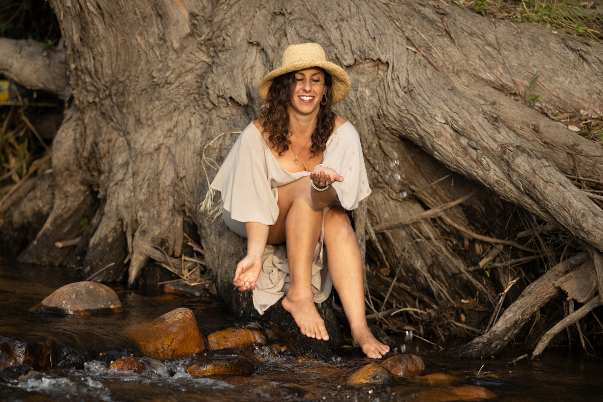 A woman sitting barefoot on rocks in a stream, wearing a wide-brimmed straw hat and a light beige dress, with a large tree root behind her.