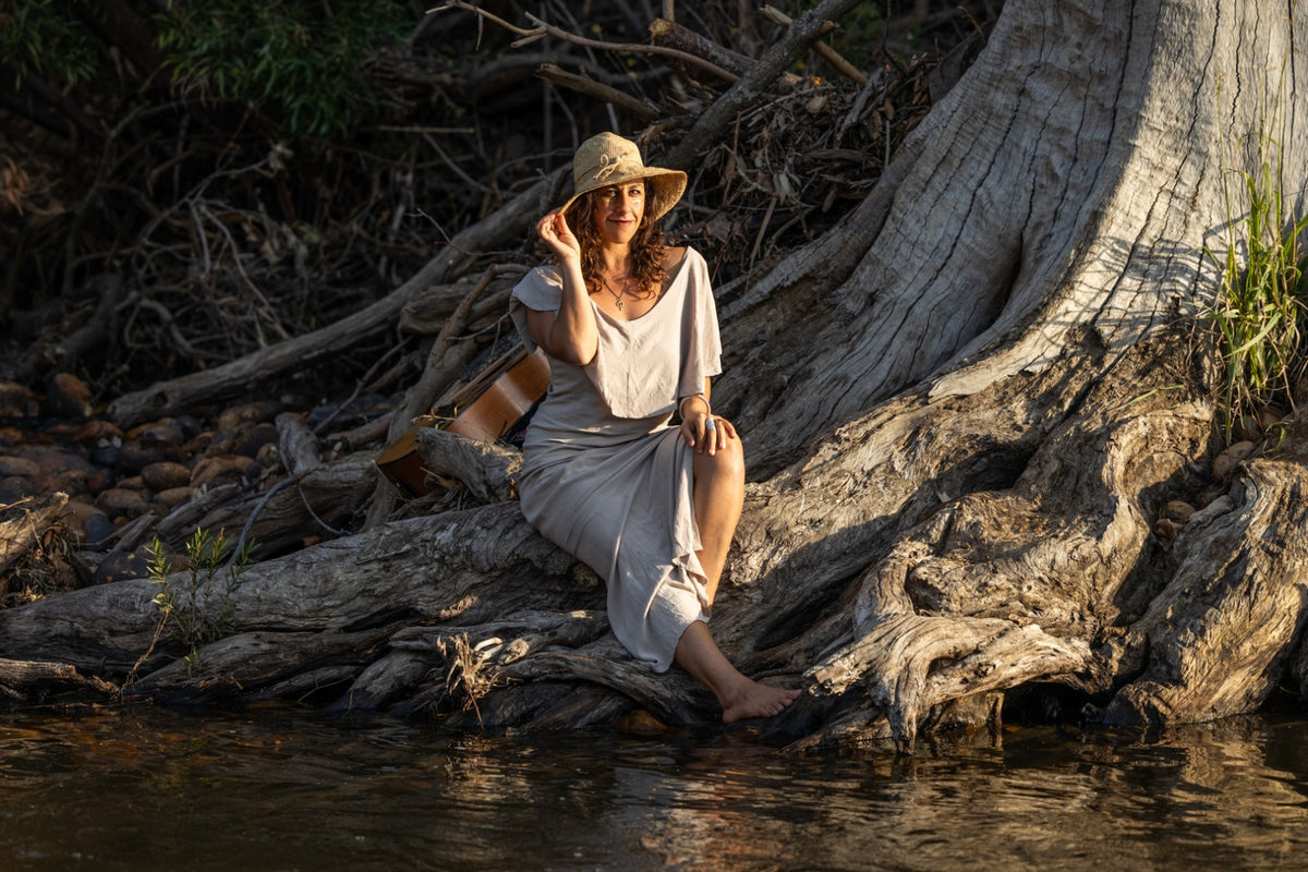 A woman wearing a beige wide-brim hat and a matching beige draped dress sits barefoot on a large tree root beside a body of water, with natural outdoor foliage and branches in the background.