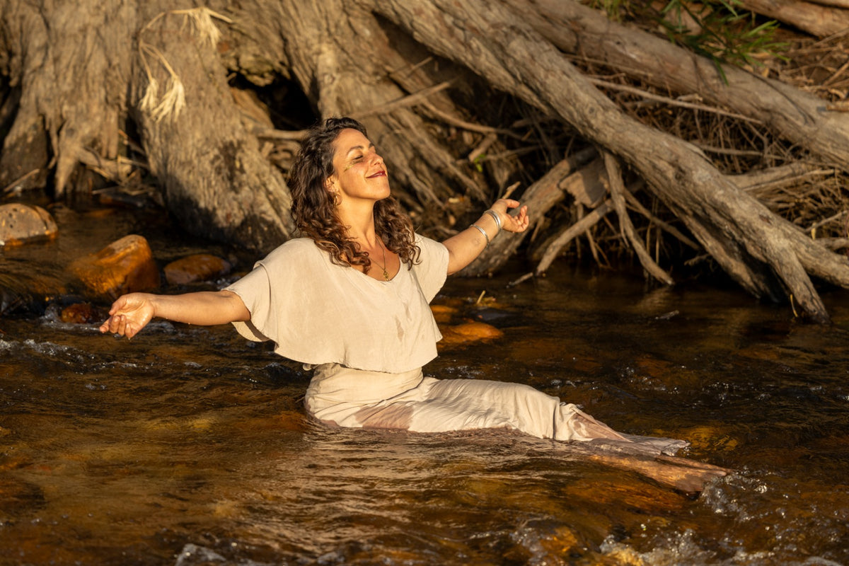 A woman sitting in a shallow stream with her arms outstretched, wearing a light beige dress, with large tree roots and rocks behind her.
