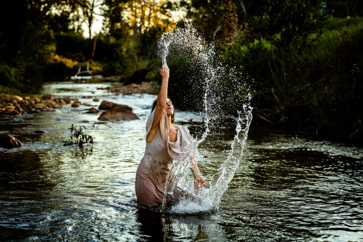 A woman in a flowing stone coloured dress stands in a shallow river, throwing water high into the air with her arm raised. The scene is set outdoors in a natural wooded stream at golden hour, with sunlight filtering through the trees and dark green foliage along the banks.