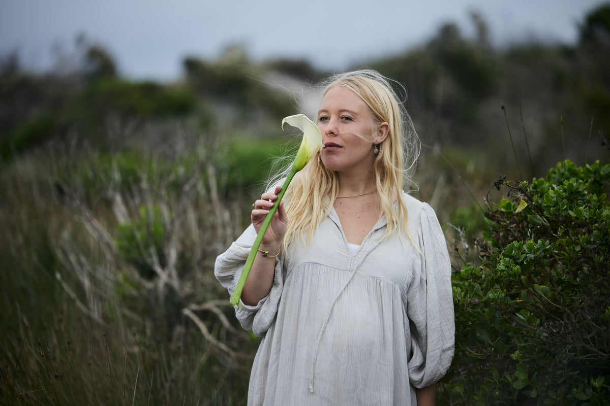 A blonde woman in a pale blue organic cotton dress stands outdoors in a grassy field, holding a white calla lily flower to her face.