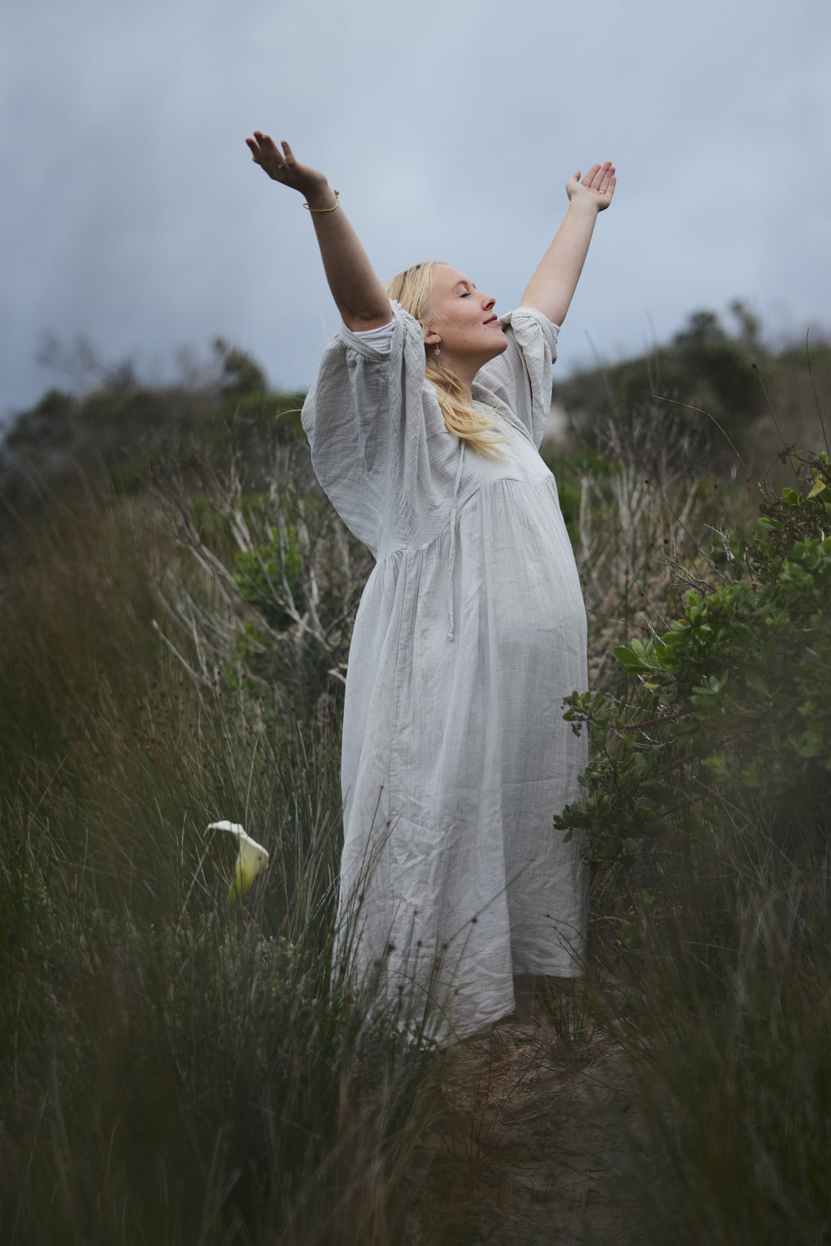 A pregnant woman in a long, flowing dress stands outdoors in a grassy field with her arms raised, looking upward under an overcast sky.