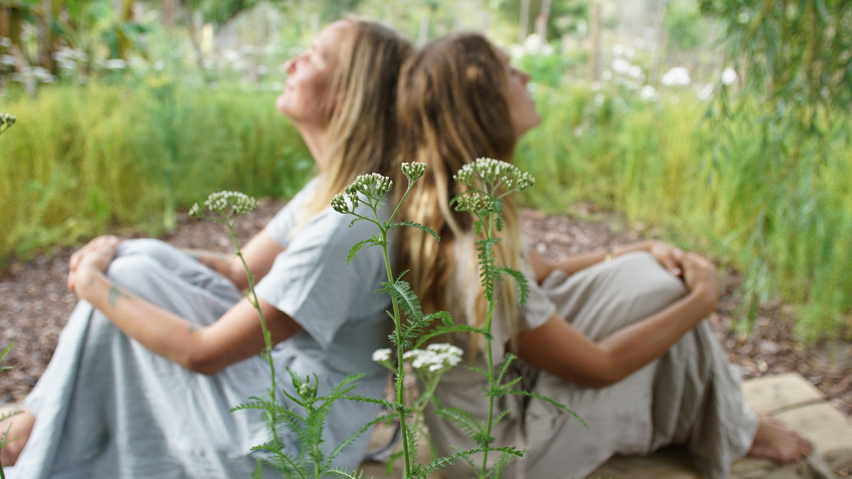 Two women sitting back-to-back in a garden, wearing soft, flowing, pale blue and stone coloured dresses. White yarrow flowers in the foreground add depth to the scene.