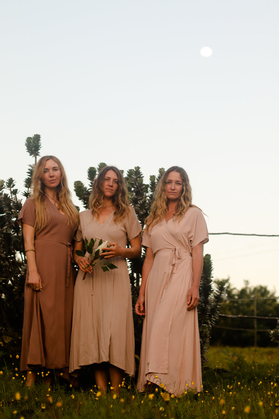 Three women standing outdoors in a grassy field at dusk, wearing matching long dresses in earthy neutral tones: one brown, one beige, and one blush pink. Trees and a pale sky with a visible moon are in the background.