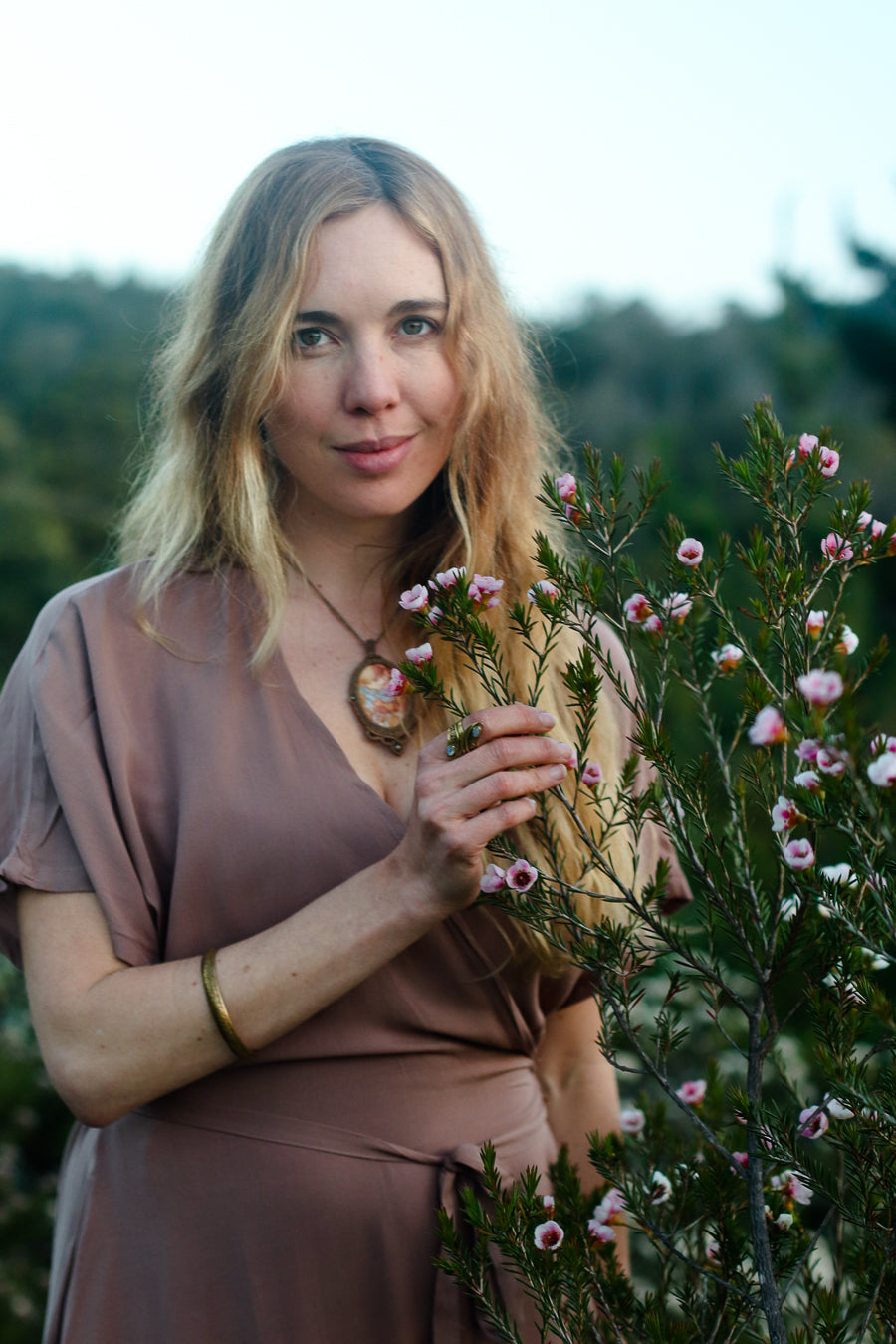 A woman with blonde hair wearing a dusty rose-brown wrap dress and a gold pendant necklace, standing outdoors in a field of pink and white flowers.