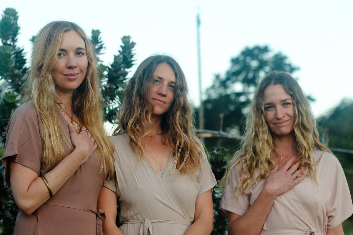Three women standing outdoors in front of trees, each wearing a soft neutral wrap dress in different shades of taupe, beige, and blush. They have their hands over their chests.