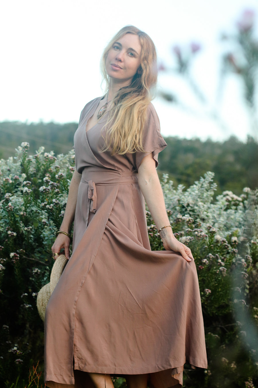 A woman wearing a dusty rose wrap midi dress stands in a field of white flowers, holding out one side of the dress to show its flow. She is holding a straw hat in her other hand.