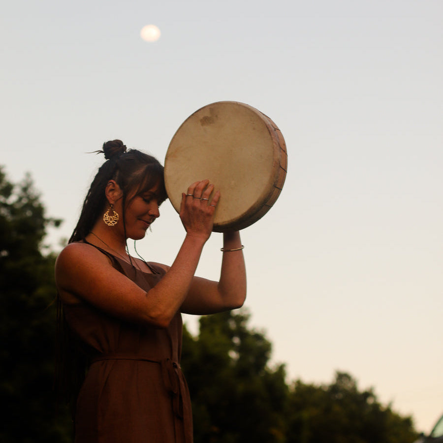 A woman in a brown linen dress holds a large hand drum overhead outdoors at sunset, with trees in the background and a visible moon in the sky.